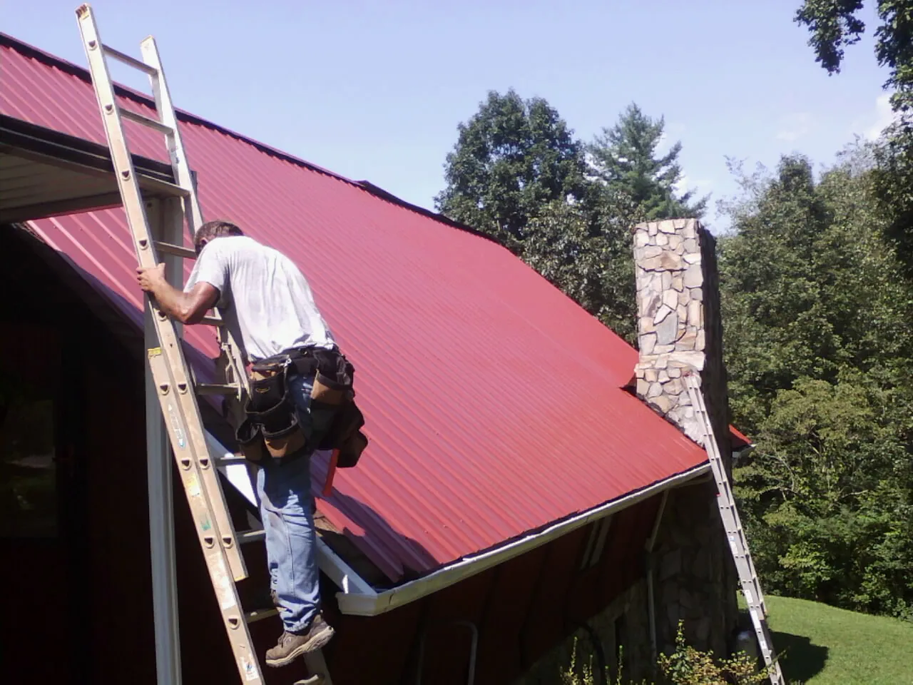 Roofer on a ladder beside a red metal roof and stone chimney — Soffit & Fascia Repair by Roofing Brothers in Mound City